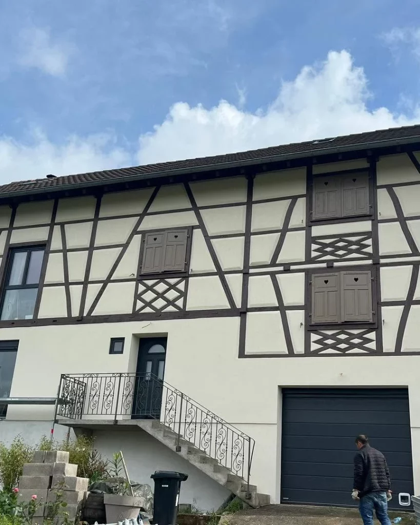 Maison à colombages après rénovation de la façade : enduit clair, colombages et volets bruns, porte de garage anthracite